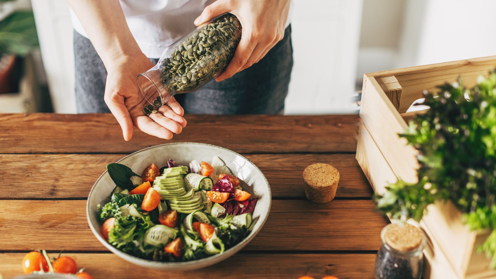 Woman&#039;s hands pouring pumpkin seeds into bowl of salad, with leaves, tomatoes, cucumber, sitting on wooden garden table, representing the best diet for longevity
