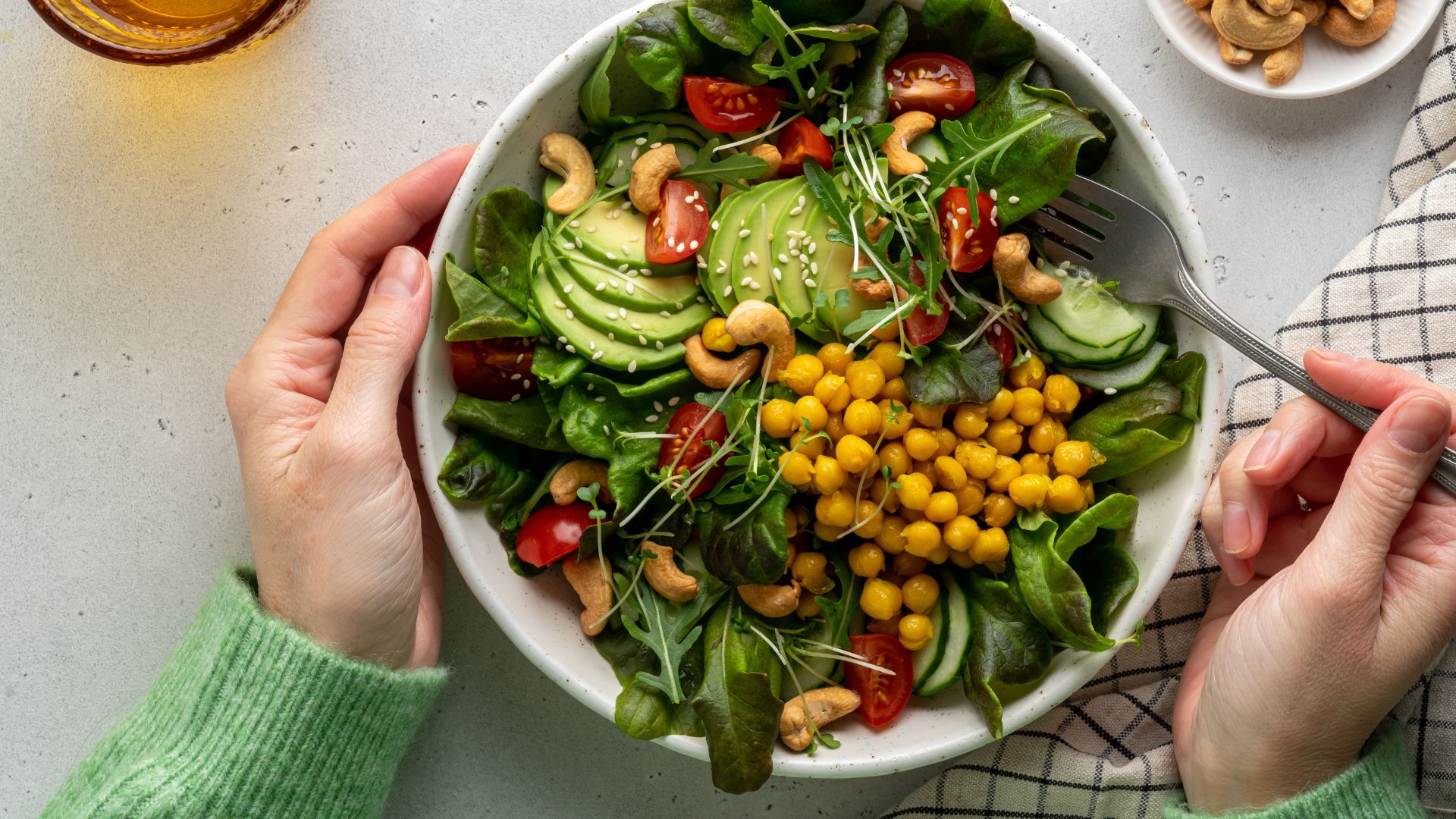 Bowl of leafy vegetables, chickpeas and fibre-rich foods with woman&#039;s hands around the bowl and tea towel on the counter to represent the process of making postbiotics